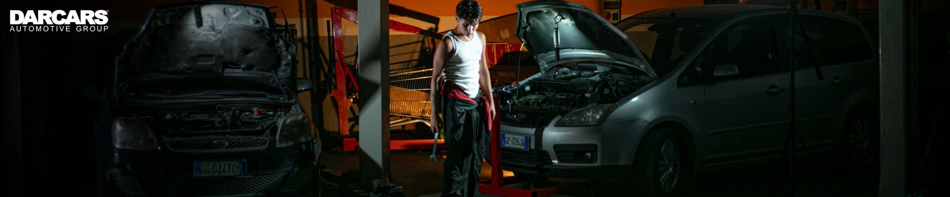 Wide shot of a mechanic standing between two cars with open hoods inside a automotive service garage.