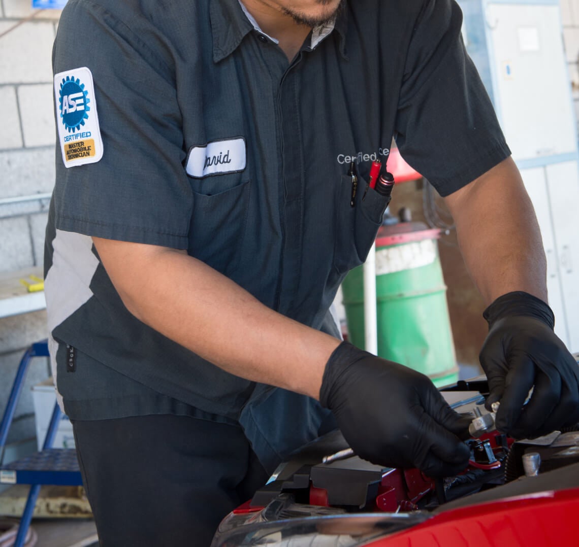 Close-up shot of an ASE Certified Technician in black gloves working on a car engine in a repair shop.