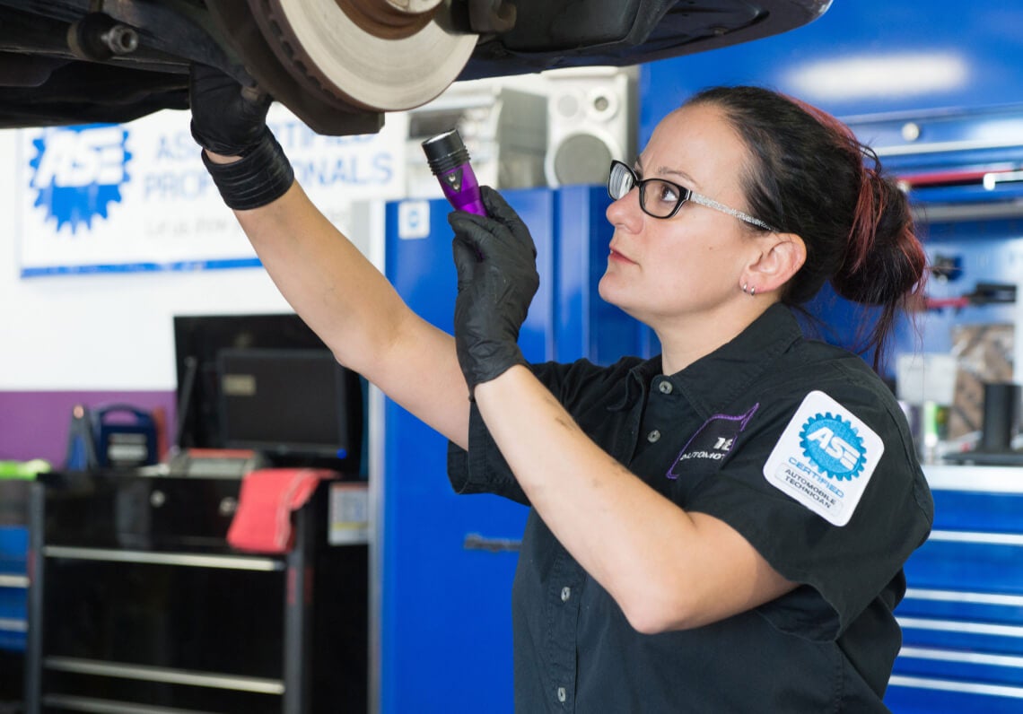 Medium shot of an ASE Certified technician using a flashlight to inspect vehicle brakes on a lift.