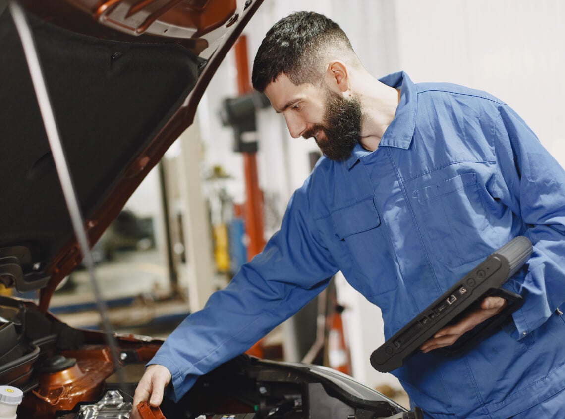 Medium shot of a mechanic in blue coveralls holding a diagnostic tablet while inspecting a car.