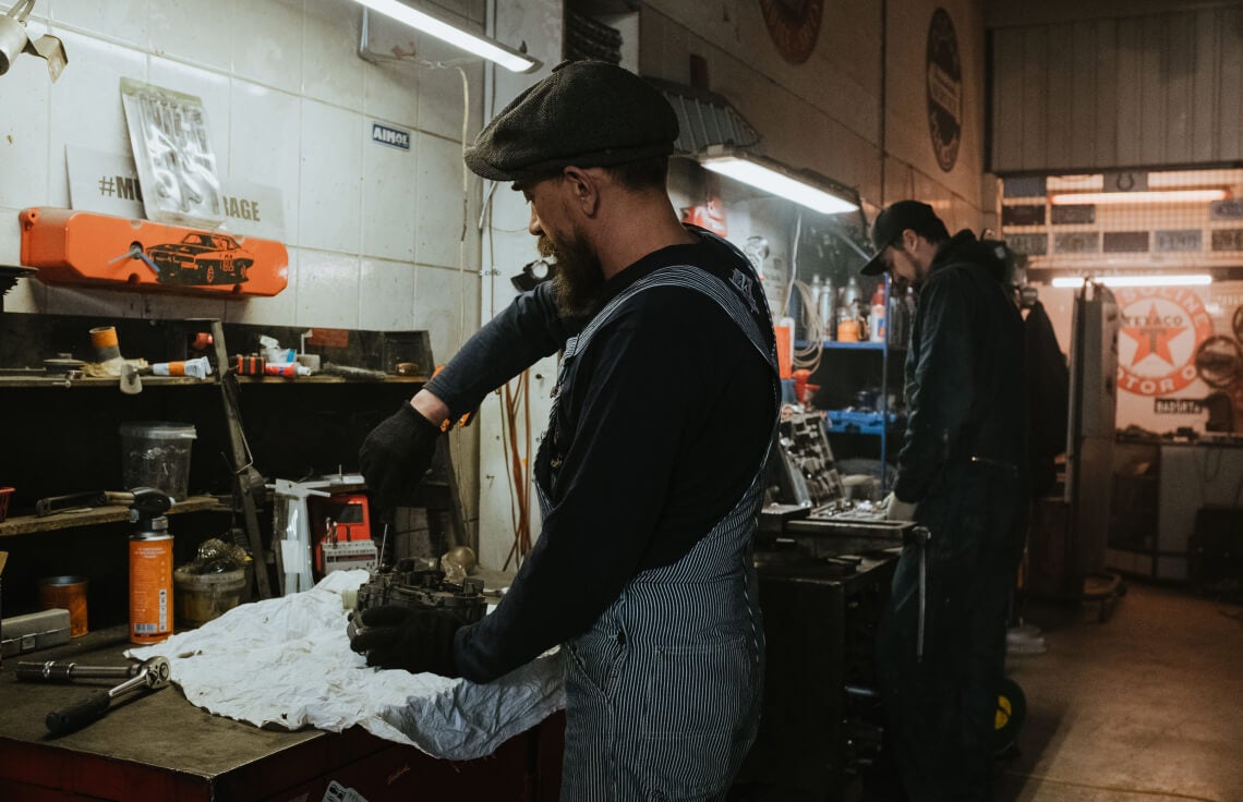 Medium shot of a bearded technician working on an engine part under bright workshop lights.