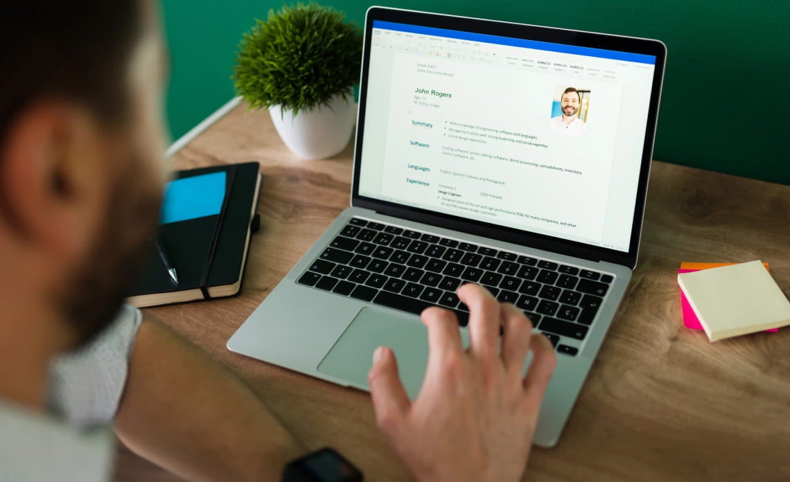 A person reviews a resume on a laptop at a wooden desk, with a notebook, pen, and sticky notes nearby. The scene conveys focus and professionalism.
