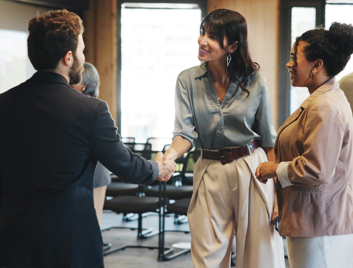 Three people in a modern office setting. A woman in a blue blouse smiles while shaking hands with a man in a suit; another woman stands nearby, observing warmly.