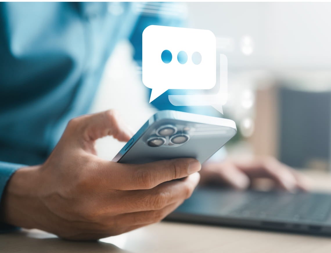 A person in a blue shirt holds a smartphone, with a chat bubble icon above it. They’re using a laptop in a bright, modern workspace.